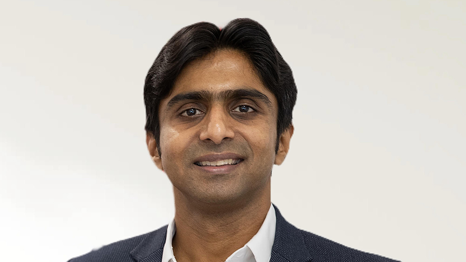 Portrait of a man wearing a navy suit jacket and white shirt, smiling against a light neutral background.